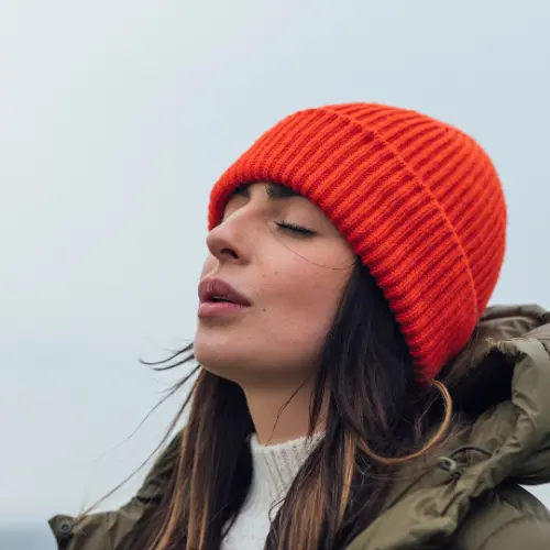 A woman wearing an orange knit beanie closes her eyes, looking serene and relaxed. She wears a warm jacket and appears to be enjoying a moment outdoors, with a soft, neutral background.