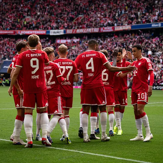 A group of FC Bayern Munich players in red and white uniforms huddles together on the field, celebrating and strategizing. The background features a cheering crowd in a stadium, creating a vibrant atmosphere during a match.