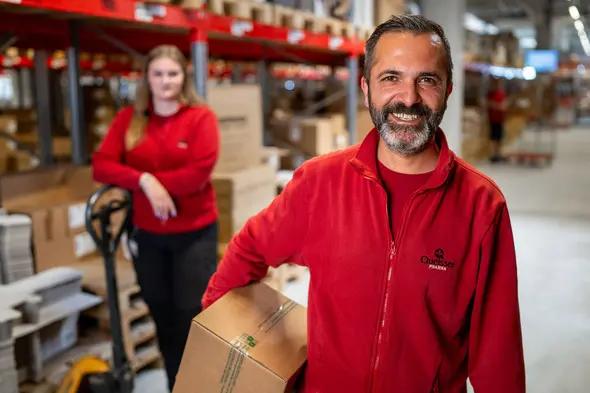 A smiling man in a red jacket holds a box in a warehouse setting. Behind him, a woman in a similar red outfit stands with her hands on her hips, surrounded by stacked boxes and shelves. The environment suggests an organized workspace.