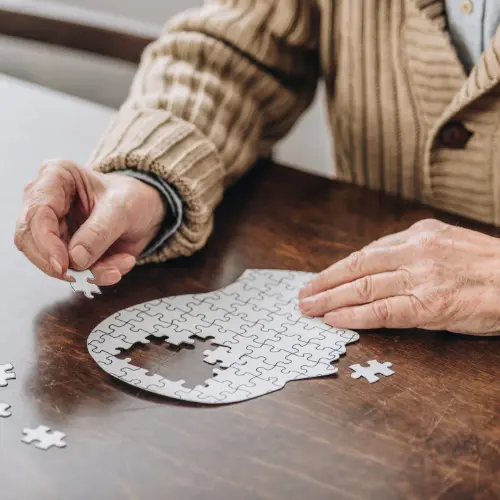 An elderly person is sitting at a table, assembling a white jigsaw puzzle piece. Their hands are positioned over a partially completed puzzle design, with additional scattered pieces nearby. The focus is on the act of connecting the puzzle.