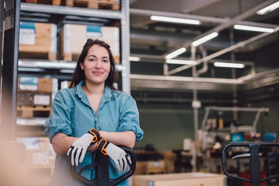 A woman wearing a denim shirt and gloves stands confidently with her arms crossed over a pallet jack in a warehouse. The background features shelves filled with boxes and various warehouse equipment.