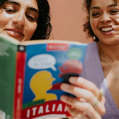 Two women are sitting close together, smiling while looking at a colorful book titled "Italian." One woman holds the book, which features playful illustrations. They appear engaged and happy, suggesting they are enjoying their time together, possibly learning or sharing something related to the book.