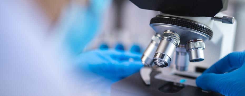A close-up view of a laboratory microscope being adjusted by a person wearing blue gloves. The focus is on the microscope's lenses and the individual's hand positioning a sample below it, suggesting a scientific or medical context.