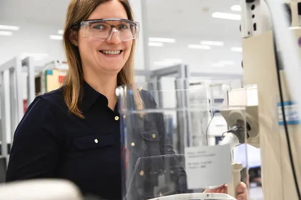 A smiling woman wearing safety glasses stands next to laboratory equipment. She appears engaged and is in a modern lab setting with shelves and other equipment in the background.
