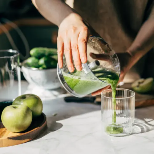 A person is pouring green juice from a blender into a clear glass. Fresh green apples and cucumbers are visible on the countertop, suggesting a healthy drink preparation. The scene is well-lit, highlighting the vibrant colors of the ingredients.