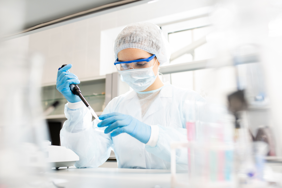 A laboratory technician in protective gear uses a pipette to measure liquid. They are wearing a lab coat, gloves, goggles, and a face mask, focused on their task in a clean and organized lab environment with laboratory equipment visible.