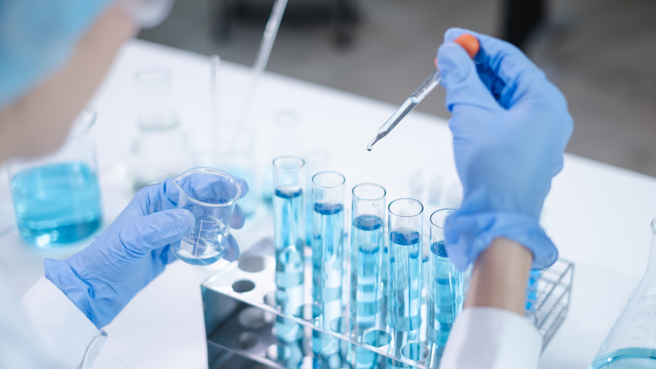 A person in a lab coat and blue gloves holds a small beaker in one hand and a dropper in the other, preparing to add liquid to a row of test tubes filled with blue liquid. Various laboratory glassware is visible in the background.