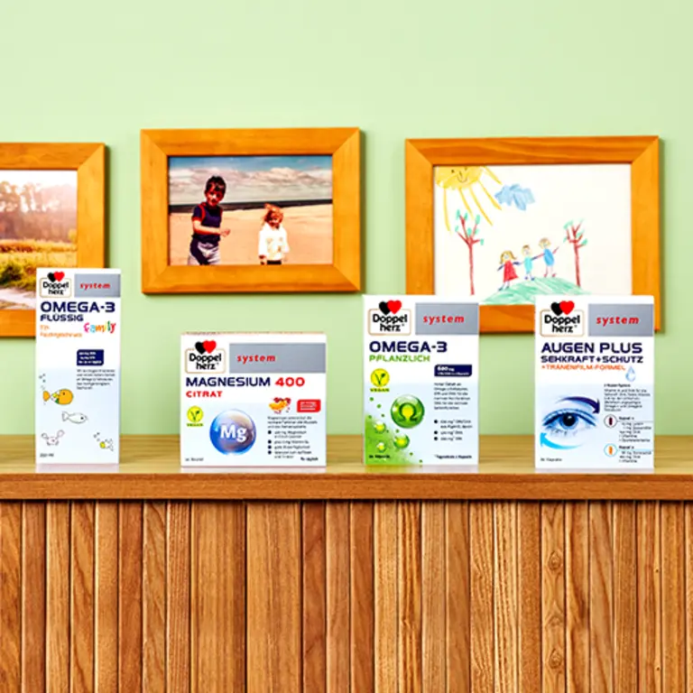 A wooden shelf displays four health product boxes against a light green wall. The products include Omega-3 capsules, Magnesium 400 Citrate, and Omega-3 plant-based, with a box labeled "Augen Plus" for eye health. Family photos and children's drawings are visible in frames above the shelf.