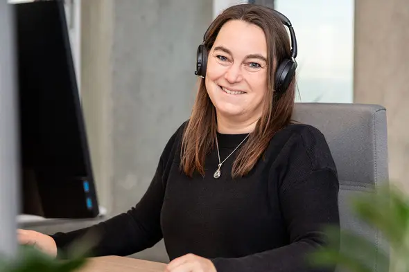 A woman wearing headphones smiles while sitting at a desk with a computer monitor. She has long, dark hair and is dressed in a black sweater. A plant is visible in the foreground. The setting appears to be an office environment.