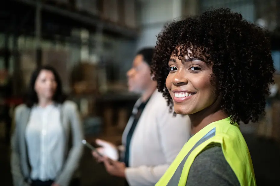 A woman wearing a bright yellow safety vest smiles at the camera, standing in a warehouse environment. Two other women are engaged in conversation in the background, with one checking her phone. The setting appears professional and industrial.