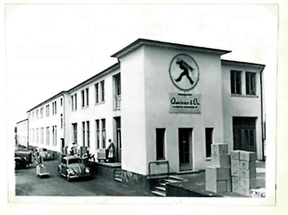A black and white photograph of a building with a sign featuring a person carrying a load. The structure has multiple windows and a staircase at the entrance. In the foreground, there are several individuals and parked vintage cars. Boxes are stacked nearby.