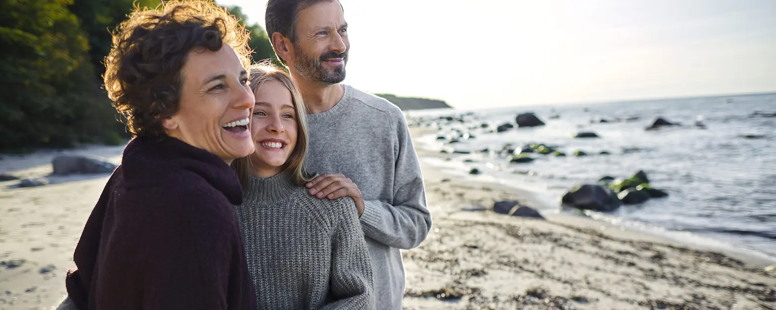 A woman and a girl smile warmly while standing close together, with a man behind them, all on a sandy beach. The sea is visible in the background, and the scene is illuminated by natural sunlight, creating a joyful and serene atmosphere.